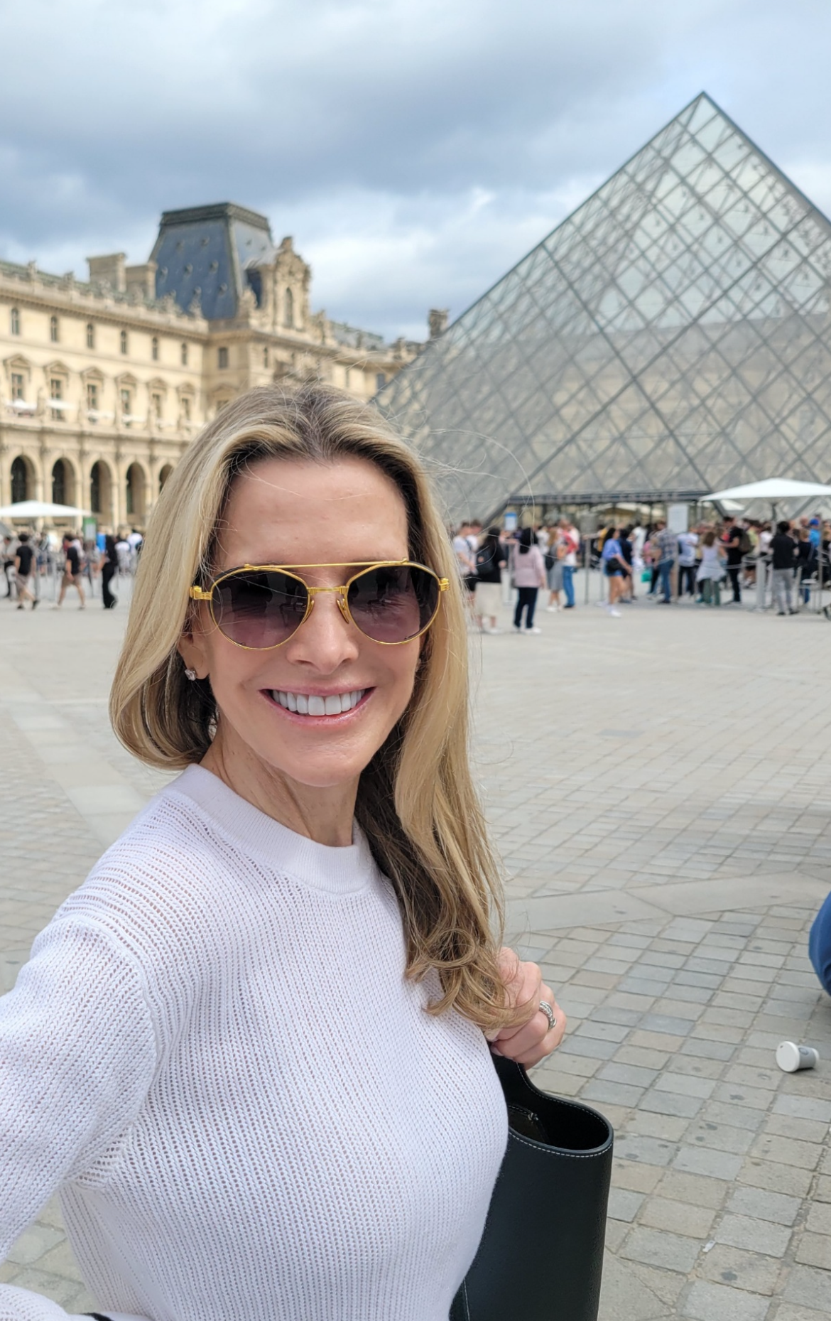 Woman in sunglasses and white sweater standing in front of the Louvre Pyramid in Paris